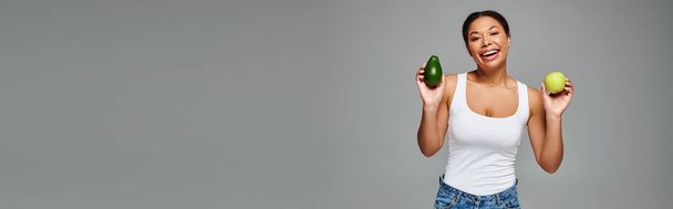 Donna afroamericana radiante con mela e avocado che promuove un'alimentazione equilibrata su grigio, striscione - Foto, immagini