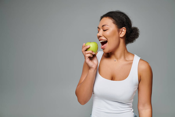 szczęśliwy Afroamerykanka kobieta z białymi zębami gryzie zielone jabłko na szarym tle, zdrowe odżywianie - Zdjęcie, obraz