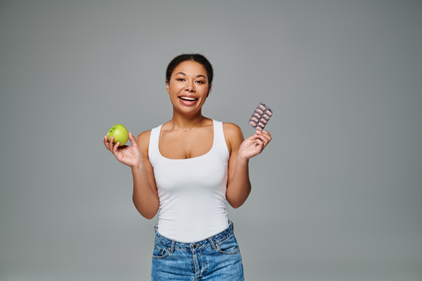 ευτυχισμένη Αφρικανή Αμερικανίδα συγκρίνει τα συμπληρώματα με πράσινο μήλο γκρι φόντο, επιλογή διατροφής - Φωτογραφία, εικόνα