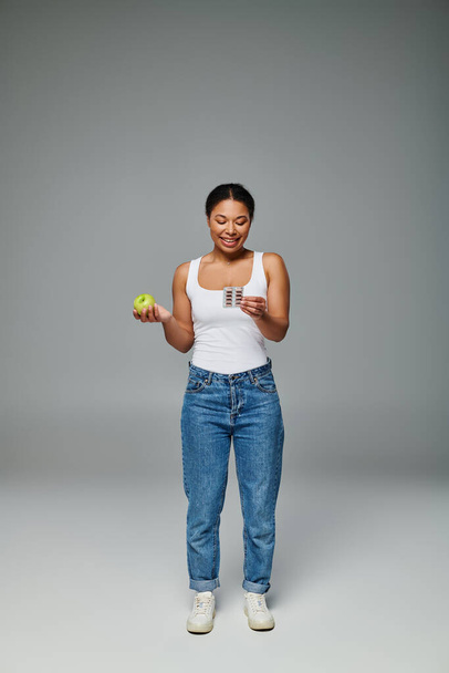счастливая африканская американка сравнивает добавки с зеленым яблоком серый фон, витамины - Фото, изображение