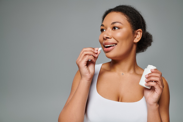mujer afroamericana feliz tomando suplementos en forma de píldora sobre fondo gris, plan dietético - Foto, imagen
