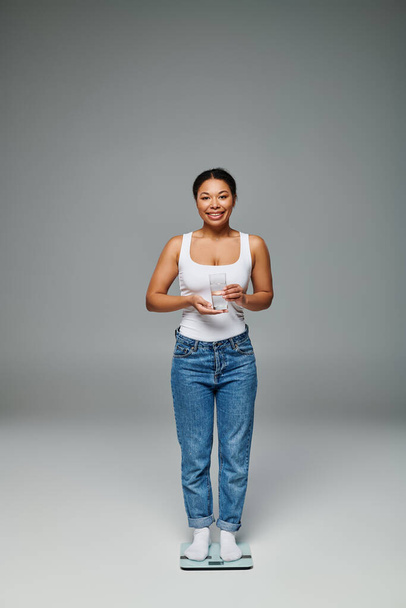 щаслива афроамериканка в повсякденному вбранні тримає склянку води і стоїть на лусках на сірому - Фото, зображення