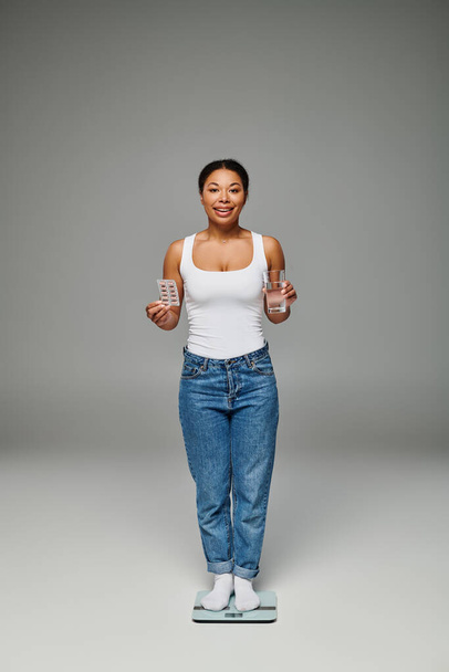 szczęśliwa Afroamerykanka trzyma szklankę wody i suplementów i stoi na skali na szarości - Zdjęcie, obraz