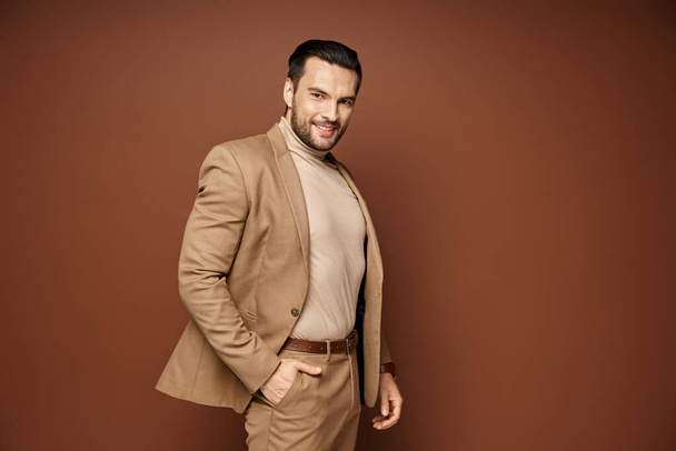 hombre alegre con cerdas posando en elegante chaqueta beige con la mano en el bolsillo sobre fondo beige - Foto, imagen