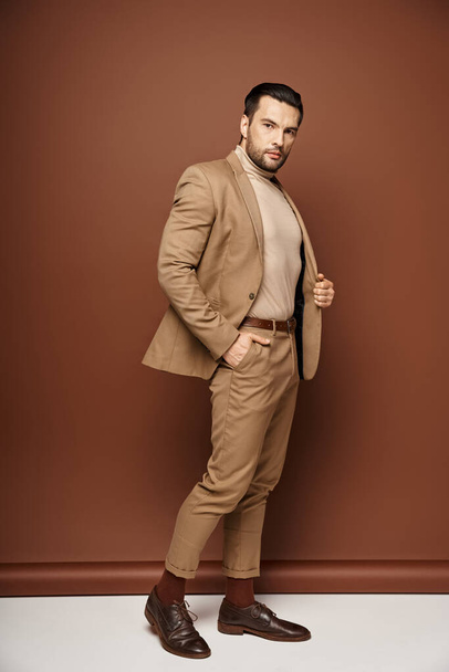 hombre suave en traje elegante mirando hacia otro lado mientras posando con la mano en el bolsillo sobre fondo beige - Foto, imagen