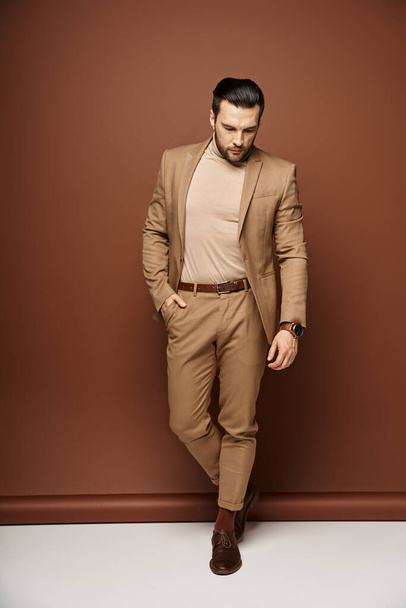 hombre guapo en traje elegante mirando hacia abajo mientras posando con la mano en el bolsillo sobre fondo beige - Foto, imagen