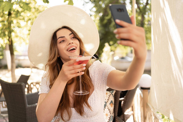 Beautiful young girl in big summer straw hat taking a selfie resting at the sunny terrace, sitting at a cafe with a cocktail. Copy space - Photo, Image