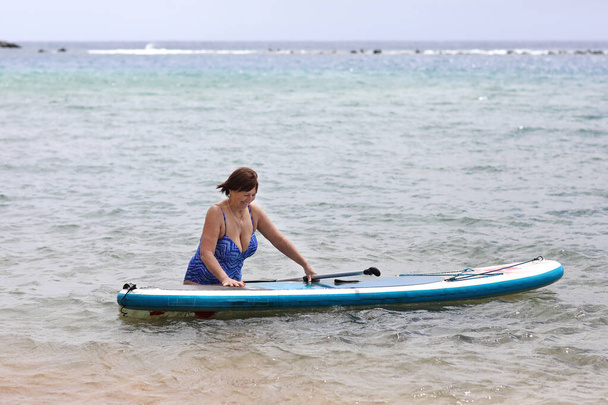 Zadní pohled dospělé ženy s nadváhou v plavkách s SUP deskou a pádlem jde do moře. Wellness žena středního věku si užívá surfování na brouzdališti na letních prázdninách. Aktivní moderní životní styl - Fotografie, Obrázek