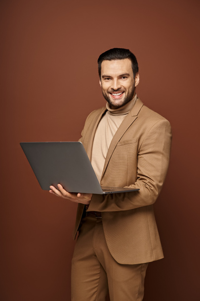 gelukkig en knap zakenman houden van zijn laptop tijdens het werken op afstand op beige achtergrond - Foto, afbeelding