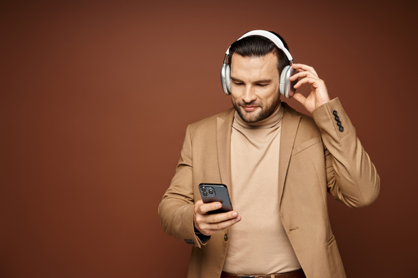 usmívající se pohledný muž v bezdrátových sluchátkách pomocí smartphonu na béžovém pozadí, digitální věk - Fotografie, Obrázek