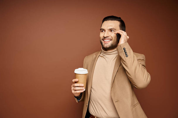 ベージュの背景にスマートフォンで電話して行くためにコーヒーを保持するスタイリッシュな服装の幸せな男 - 写真・画像