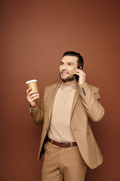 περιεχόμενο άνθρωπος σε κομψή ενδυμασία κρατώντας καφέ για να πάει και έχοντας κλήση στο smartphone σε μπεζ φόντο - Φωτογραφία, εικόνα