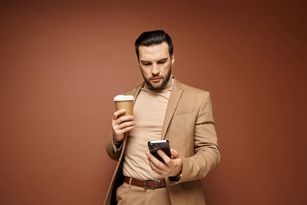 dobře vypadající muž ve stylovém oblečení drží kávu jít a pomocí svého smartphonu na béžové pozadí - Fotografie, Obrázek