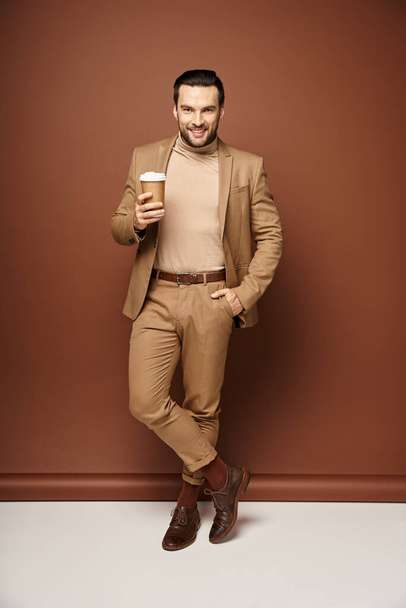 zadowolony człowiek w stylowym stroju trzymając kawę iść i pozowanie z ręką w kieszeni na beżowym tle - Zdjęcie, obraz