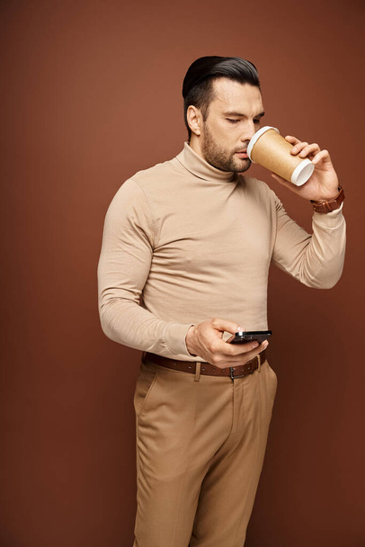 handsome man in turtleneck drinking coffee and using smartphone on beige background, social media - Photo, Image