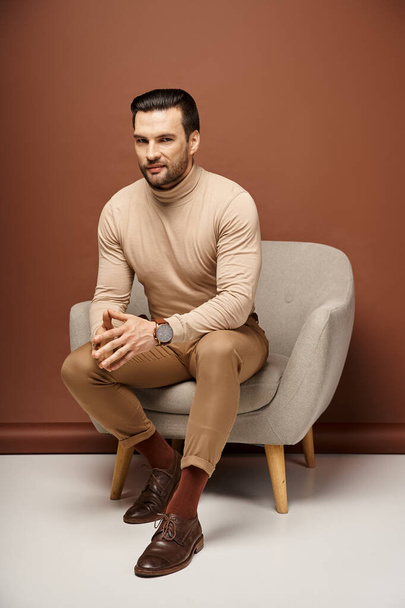 hombre guapo y astuto con cerdas sentado en cómodo sillón sobre fondo beige - Foto, imagen