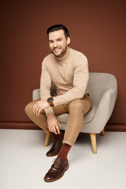 vrolijk en knap man in coltrui zittend op comfortabele fauteuil op beige achtergrond - Foto, afbeelding