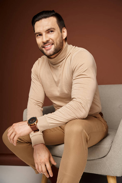 hombre positivo y guapo en cuello alto sentado en cómodo sillón sobre fondo beige - Foto, imagen