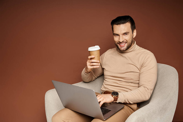 vidám ember használja laptop és a kezében kávé menni, miközben ül a karosszék bézs háttér - Fotó, kép
