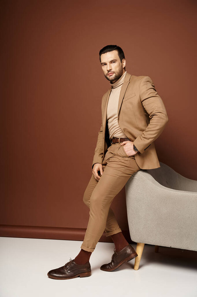 schöner Mann im eleganten Anzug posiert mit der Hand in der Tasche in der Nähe eines Sessels auf beigem Hintergrund - Foto, Bild