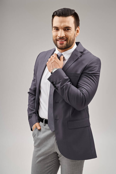 hombre feliz y guapo en traje elegante ajuste corbata y posando con la mano en el bolsillo sobre fondo gris - Foto, Imagen