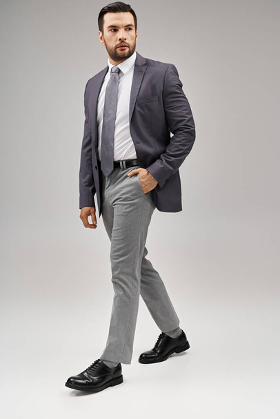 hombre guapo en traje afilado y elegante posando con la mano en el bolsillo sobre fondo gris, elegante - Foto, imagen
