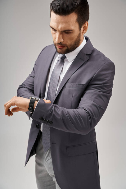 bell'uomo d'affari in posa in un abito elegante controllando il suo orologio da polso su sfondo grigio, eleganza - Foto, immagini