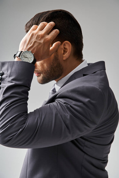 pohledný muž ve formálním oděvu s hodinkami upravující si vlasy na šedém pozadí, firemní móda - Fotografie, Obrázek