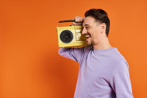 fröhlicher Mann in lila Sweatshirt mit Retro-Boombox und Musik auf orangefarbenem Hintergrund - Foto, Bild