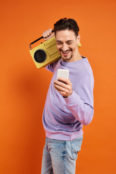 hombre alegre y barbudo usando smartphone y sosteniendo boombox retro sobre fondo naranja, música - Foto, imagen
