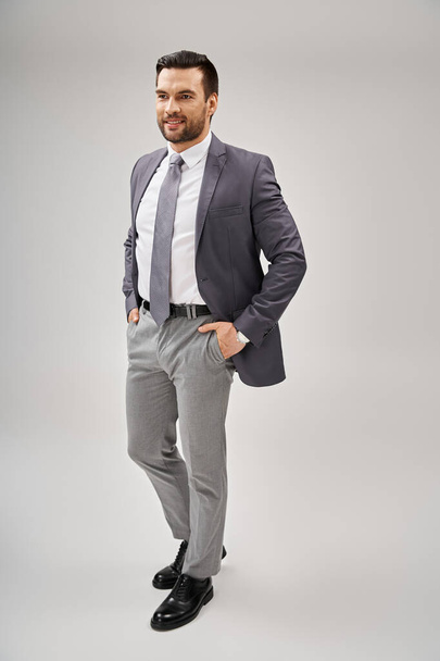portrait of happy businessman in stylish attire posing with hands in pockets on grey background - Photo, Image
