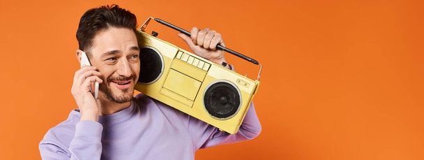 hombre alegre y barbudo hablando en el teléfono inteligente y sosteniendo boombox retro sobre fondo naranja, bandera - Foto, Imagen