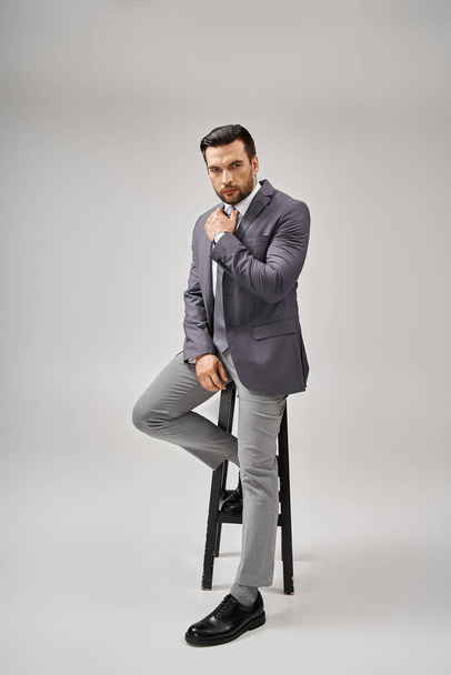gutaussehender und selbstbewusster Geschäftsmann in formaler Kleidung, angelehnt an einen hohen Hocker auf grauem Hintergrund, Eleganz - Foto, Bild