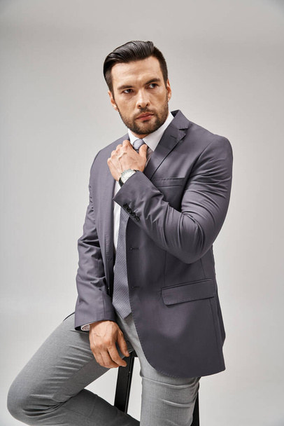 gutaussehender und selbstbewusster Geschäftsmann in formeller Kleidung, der auf einem hohen Hocker auf grauem Hintergrund lehnt - Foto, Bild