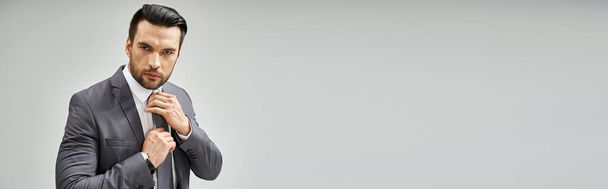вдумливий чоловік у формальному носінні регулюючи краватку на сірому фоні, корпоративний банер моди - Фото, зображення