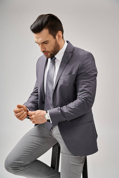 gutaussehender und selbstbewusster Geschäftsmann im Anzug, der auf einem hohen Hocker auf grauem Hintergrund sitzt, Führung - Foto, Bild