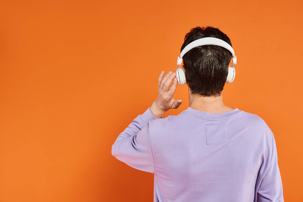 achteraanzicht van de mens in paarse trui en draadloze hoofdtelefoon luisterend naar muziek op oranje achtergrond - Foto, afbeelding