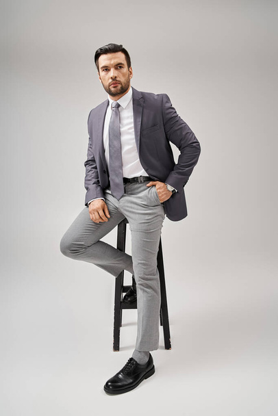 schöner Geschäftsmann mit Borsten posiert mit der Hand in der Tasche im Anzug und sitzt auf einem hohen Hocker - Foto, Bild