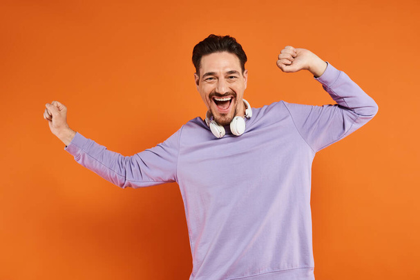 homem alegre em camisola roxa com fones de ouvido sem fio posando no fundo laranja, amante da música - Foto, Imagem