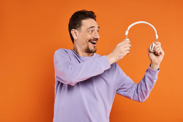 homme positif avec barbe en pull violet tenant écouteurs sans fil sur fond orange - Photo, image