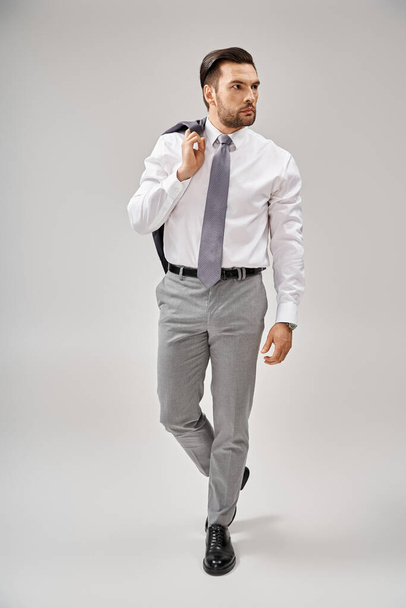 obchodník ve formálním oblečení drží bundu přes rameno při chůzi na šedém pozadí - Fotografie, Obrázek