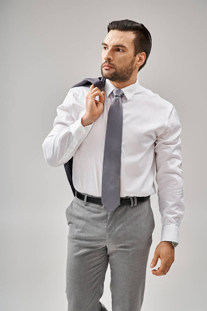 hombre de negocios de unos 30 años con una chaqueta de cerdas sobre el hombro mientras está de pie sobre un fondo gris - Foto, Imagen
