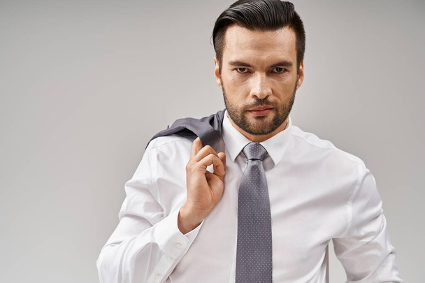 портрет бизнесмена в возрасте 30 лет с щетиной, держащей куртку через плечо, стоя на сером - Фото, изображение