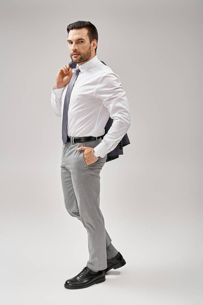 бизнесмен за 30 с щетиной, держащий куртку через плечо, стоя с рукой в кармане - Фото, изображение