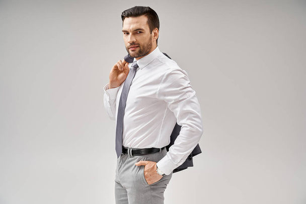 hombre de negocios con cerdas sosteniendo la chaqueta sobre el hombro mientras está de pie con la mano en el bolsillo en gris - Foto, Imagen