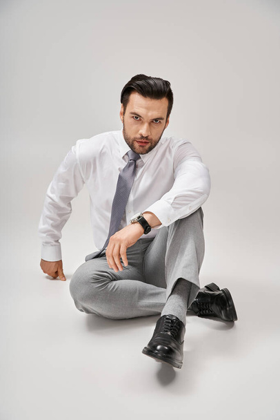 Hombre de negocios carismático y guapo en ropa formal sentado en el suelo en el estudio con fondo gris - Foto, imagen