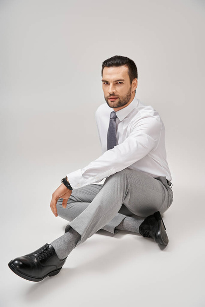 knappe zakenman in elegante formele kleding zittend op de vloer in studio met grijze achtergrond - Foto, afbeelding