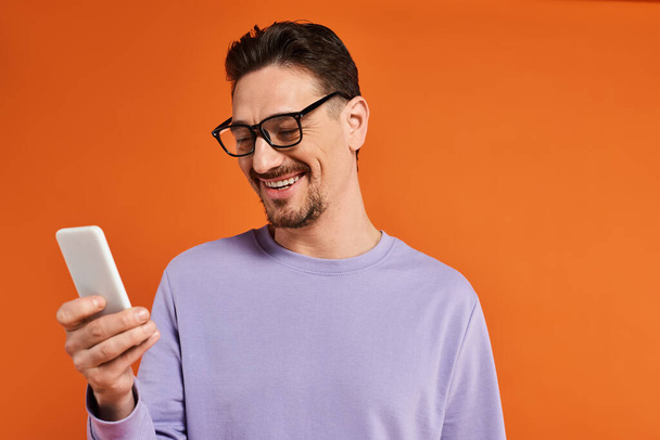 χαρούμενος άνθρωπος με γυαλιά ηλίου και μωβ πουλόβερ χρησιμοποιώντας smartphone σε πορτοκαλί φόντο, γραπτών μηνυμάτων - Φωτογραφία, εικόνα