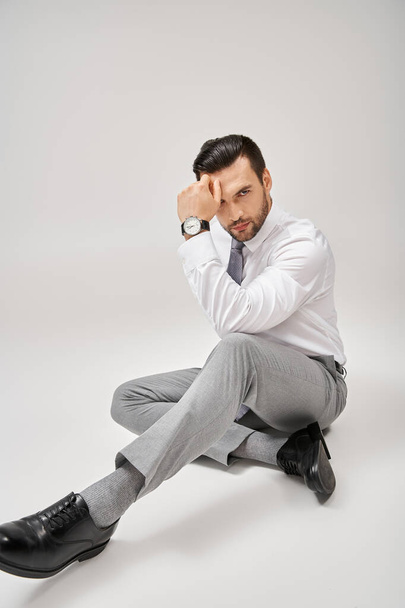 stijlvolle en charmante zakenman in formele kleding zittend op de vloer in studio met grijze achtergrond - Foto, afbeelding