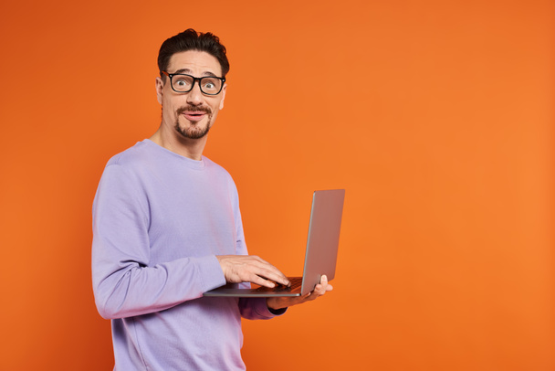 オレンジ色の背景のラップトップを使用して眼鏡と紫色のセーターで驚いた男,リモートワーク - 写真・画像